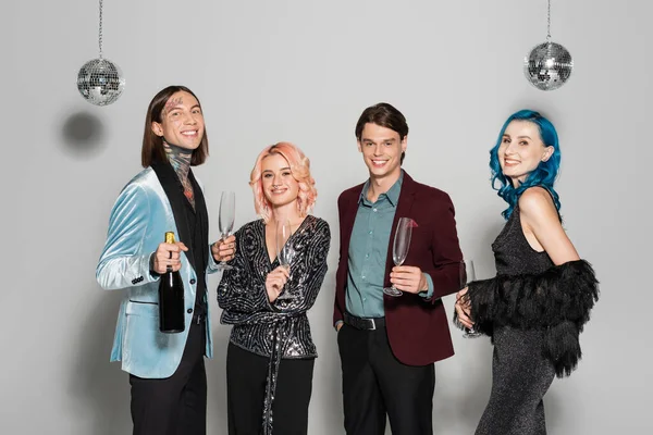 Elegant nonbinary friends with champagne smiling at camera near disco balls on grey background — Stock Photo