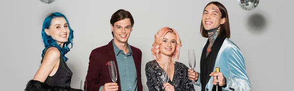 Cheerful queer people with champagne glasses smiling at camera during christmas party on grey background, banner — Stock Photo