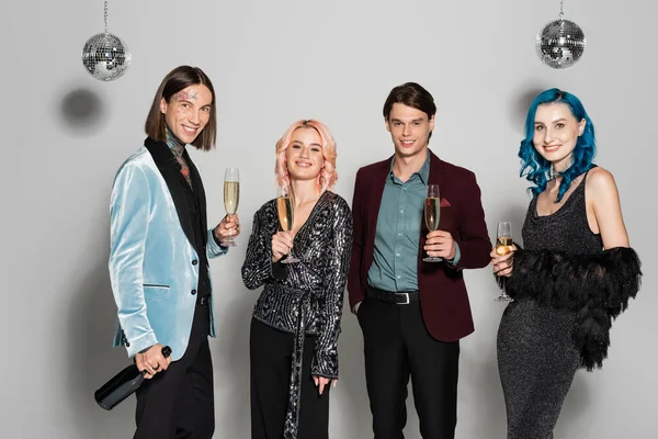 Smiling nonbinary friends in elegant festive clothes holding champagne during new year party on grey background — Stock Photo