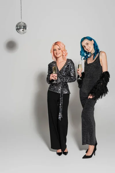 Full length of stylish nonbinary friends with champagne glasses smiling at camera on grey background — Stock Photo