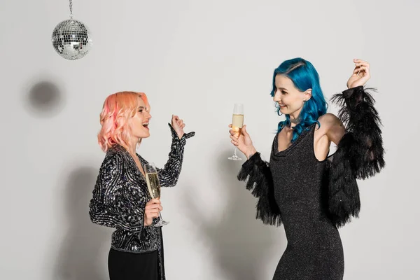 Vue latérale d'amis non binaires élégants et excités avec des verres à champagne dansant pendant la fête de Noël sur fond gris — Photo de stock