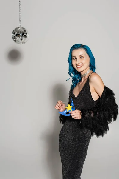 Elegant woman with blue hair holding party mask and smiling at camera near disco ball on grey background — Stock Photo