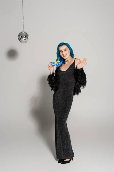 Volle Länge der lächelnden Frau in schwarzem eleganten Kleid mit Party-Maske und winkender Hand auf grauem Hintergrund — Stockfoto