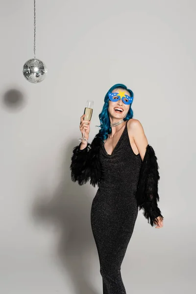 Lachende Frau mit blauer Perücke und Partymaske, die Champagner in der Hand hält und vor grauem Hintergrund wegschaut — Stockfoto