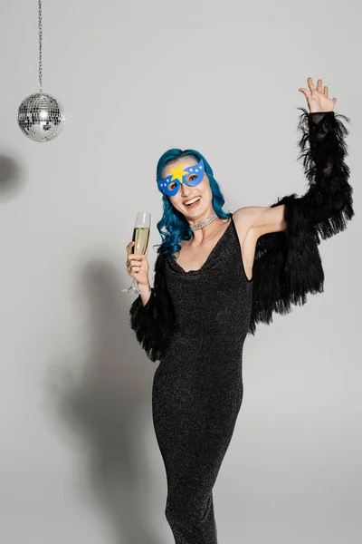 Fröhliche Frau in Partymaske und schwarzer Kunstpelzjacke mit Champagner in der Hand und winkender Hand auf grauem Hintergrund — Stockfoto