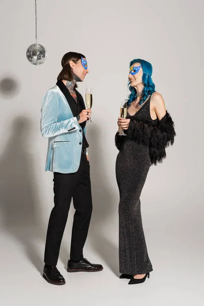 Piena lunghezza di eleganti amici queer in maschere partito che tengono bicchieri di champagne su sfondo grigio — Foto stock