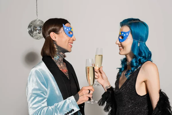 Vista lateral de elegantes amigos no binarios en máscaras de fiesta tintineo copas de champán sobre fondo gris - foto de stock