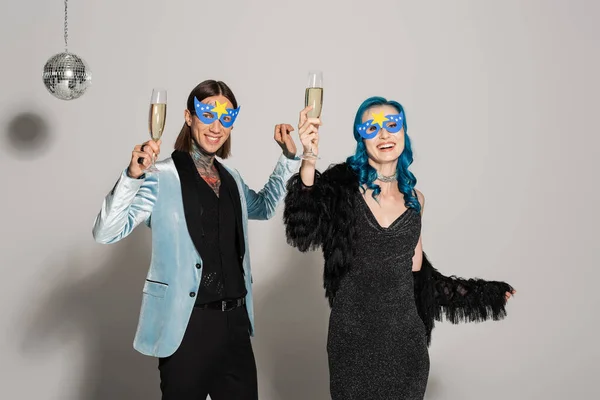 Stylish nonbinary friends in party masks toasting with champagne while smiling at camera on grey background — Stock Photo