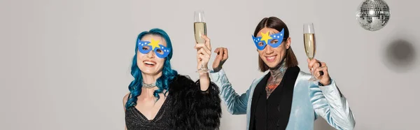 Счастливые небинарные друзья в масках для вечеринок, держащие бокалы для шампанского и смотрящие в камеру на сером фоне, баннер — стоковое фото