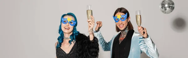 Happy nonbinary friends in party masks holding champagne glasses and looking at camera on grey background, banner — Stock Photo