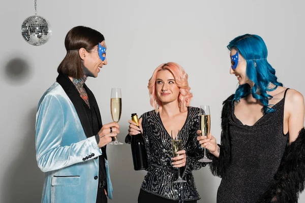 Queer person holding champagne bottle near nonbinary friends in party masks on grey background — Stock Photo