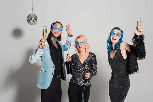 Tattooed nonbinary person holding champagne glass and waving hand near nonbinary friends in party masks on grey background — Stock Photo