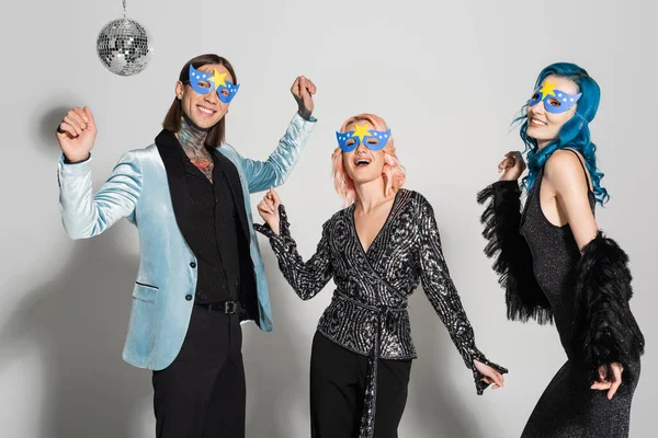 Щасливі квір-люди в елегантному одязі та вечірніх масках танцюють під час новорічної вечірки на сірому фоні — стокове фото