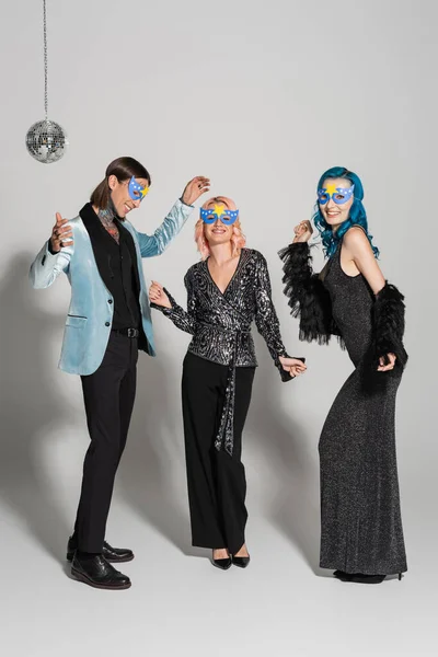 Full length of nonbinary friends in festive clothes and party masks dancing on grey background — Stock Photo