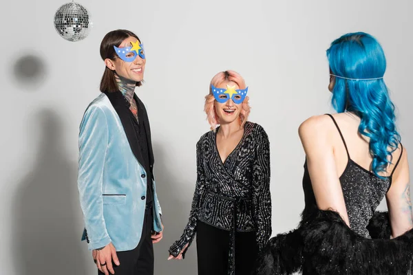 Cheerful queer friends in elegant clothes and party masks smiling at each other on grey background — Stock Photo