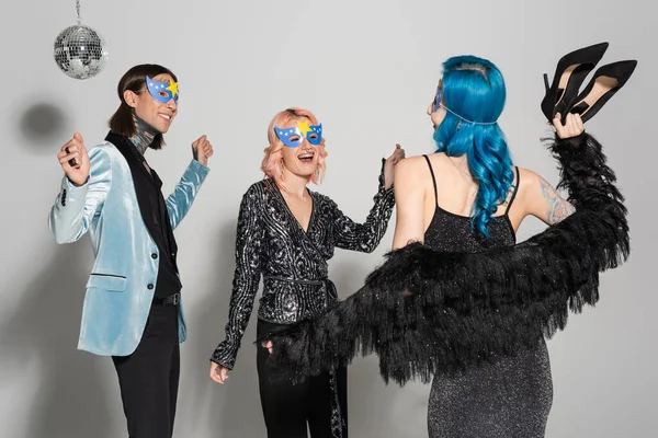 Queer person in blue wig holding high heeled shoes while dancing with nonbinary friends on grey background — Stock Photo