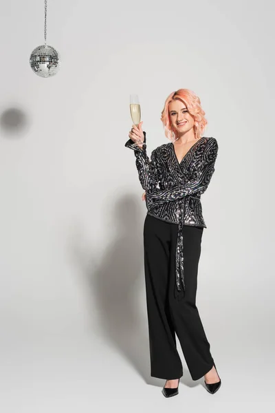 Pleine longueur de femme en chemisier brillant et pantalon noir debout avec verre de champagne sur fond gris — Photo de stock