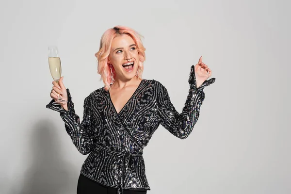 Erstaunte und stilvolle Frau mit Champagnerglas, die wegschaut und mit dem Finger auf grauen Hintergrund zeigt — Stockfoto