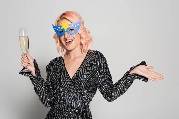 Elegant woman in party mask holding champagne glass and pointing with hand isolated on grey — Stock Photo