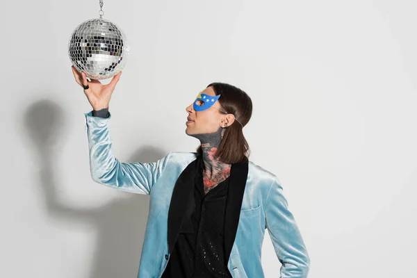 Queer personne en élégant blazer de soie et masque de fête touchant boule disco brillant sur fond gris — Photo de stock