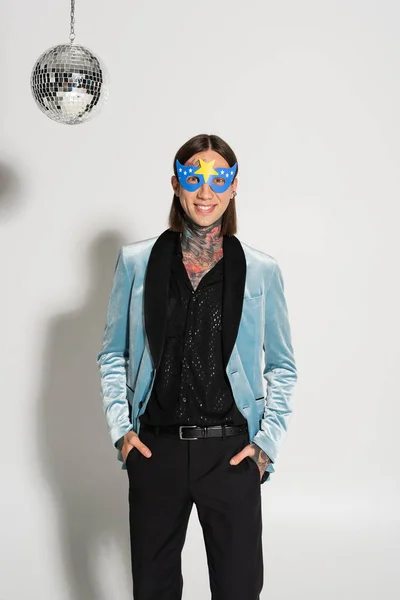 Cheerful tattooed nonbinary person in party mask standing with hands in pockets on grey background — Stock Photo