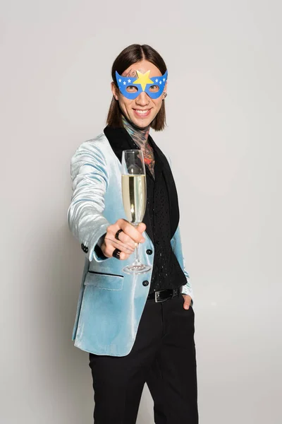 Queer persona en partido máscara y seda chaqueta celebración champán y sonriendo a la cámara en gris fondo - foto de stock
