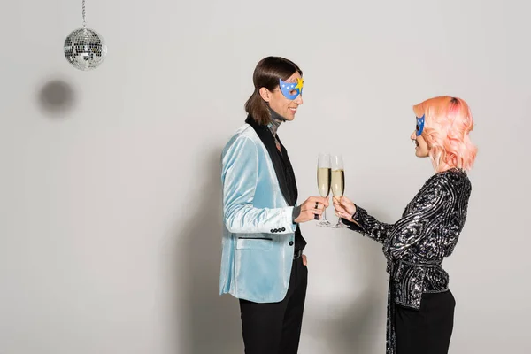 Vue latérale de heureux amis non binaires dans des masques de fête griller avec du champagne près de bal disco sur fond gris — Photo de stock