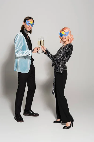 Comprimento total de amigos gays elegantes em máscaras de festa batendo copos de champanhe e sorrindo para a câmera no fundo cinza — Fotografia de Stock
