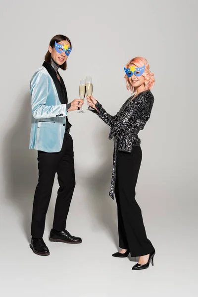 Pleine longueur d'amis queer élégants masques de fête cliquetis verres à champagne et souriant à la caméra sur fond gris — Photo de stock