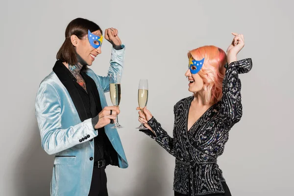 Personas queer elegantes y felices en máscaras de fiesta bailando con copas de champán sobre fondo gris - foto de stock