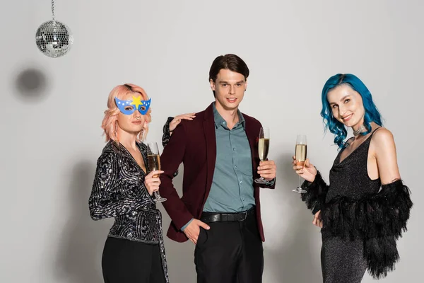 Elegante queere Freunde mit Champagnergläsern, die in die Kamera lächeln, in der Nähe einer nichtbinären Person, die mit der Hand in der Tasche auf grauem Hintergrund steht — Stockfoto