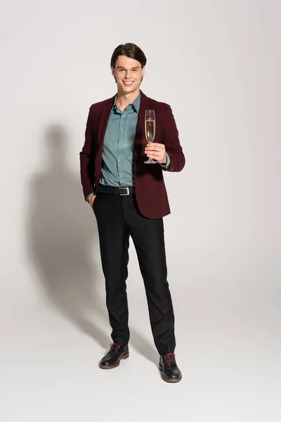 Piena lunghezza di uomo sorridente con bicchiere di champagne in piedi con mano in tasca di pantaloni neri su sfondo grigio — Foto stock