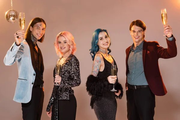 Joyful nonbinary people in stylish clothes toasting with champagne and looking at camera on beige background — Stock Photo