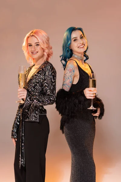 Queer friends in festive elegant clothes holding champagne and smiling at camera on beige background — Stock Photo