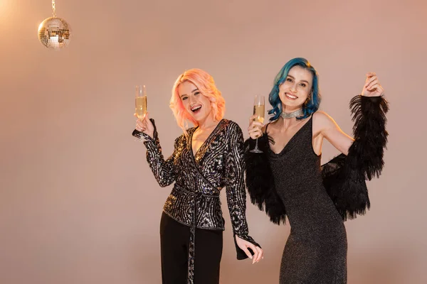 Amigos no binarios emocionados y elegantes sonriendo a la cámara y posando con copas de champán sobre fondo beige - foto de stock