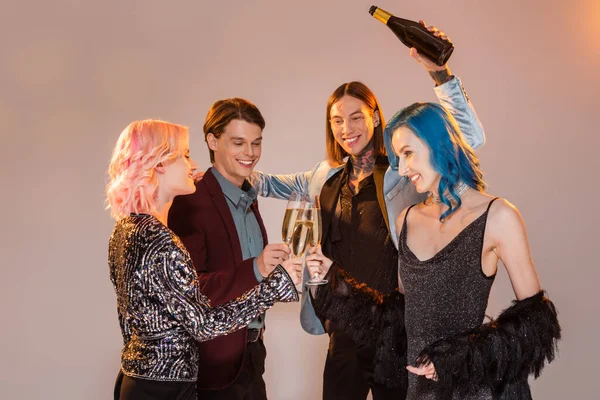 Tattooed nonbinary person holding champagne bottle near queer friends clinking glasses on grey background — Stock Photo