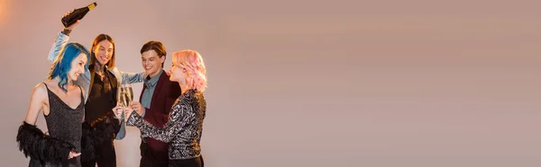 Tattooed queer person holding champagne bottle near nonbinary friends clinking glasses on grey background, banner — Stock Photo