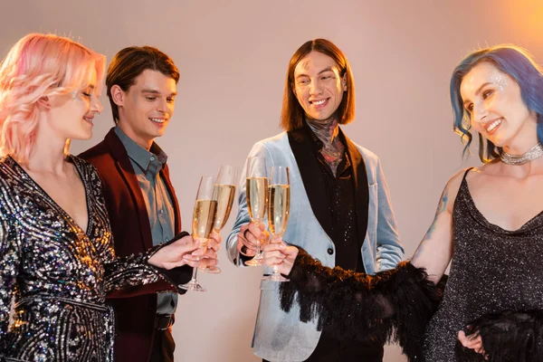 Sonriente queer personas en ropa de moda tintineo copas de champán sobre fondo gris - foto de stock