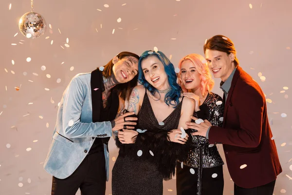 Feliz e elegante queer amigos abraçando e olhando para câmera sob queda confetti no fundo bege — Fotografia de Stock