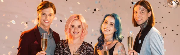 Cheerful nonbinary friends with champagne glasses looking at camera under falling confetti on beige background, banner — Stock Photo