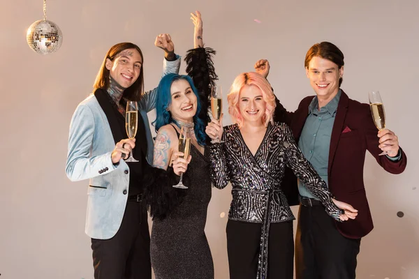 Cheerful nonbinary people with champagne glasses and raised hands on beige background — Stock Photo