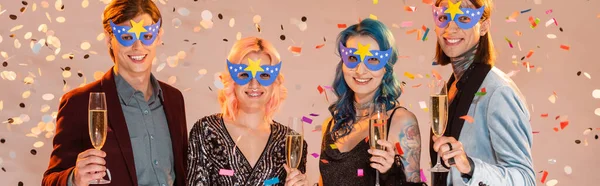 Smiling queer friends in party masks holding champagne glasses under falling confetti on beige background, banner — Stock Photo