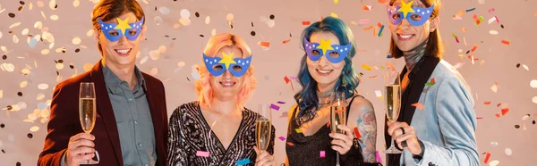 Sonrientes amigos queer en máscaras de fiesta sosteniendo copas de champán bajo la caída de confeti sobre fondo beige, bandera - foto de stock