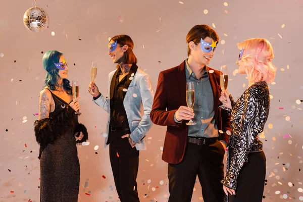 Happy queer people in party masks holding champagne glasses and talking under falling confetti on beige background — Stock Photo