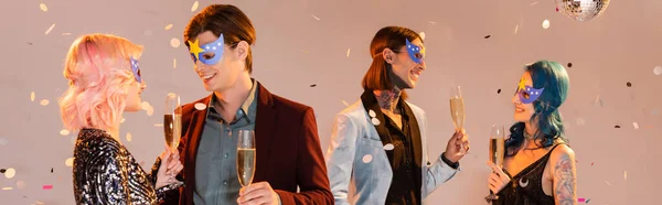 Cheerful nonbinary people talking while standing with champagne glasses under falling confetti on beige background, banner — Stock Photo