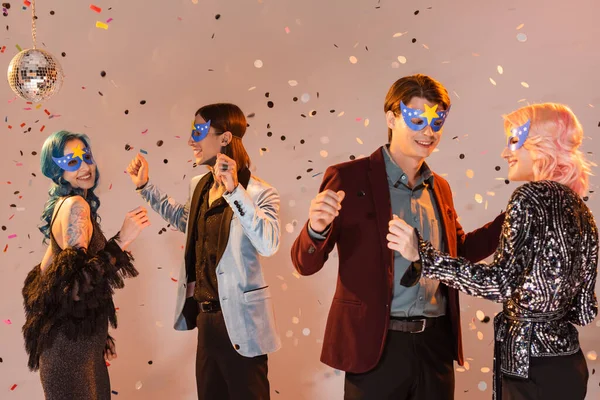 Amigos queer de moda en máscaras de fiesta bailando bajo la caída de confeti mientras se celebra la Navidad sobre fondo beige - foto de stock