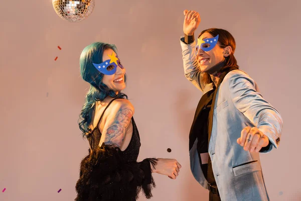 Aufgeregte queere Freunde in stylischer Kleidung und Partymasken, die auf beigem Hintergrund tanzen — Stockfoto