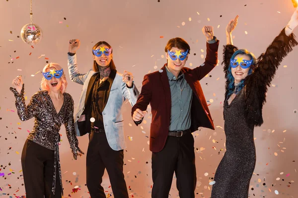 Возбужденные голубые друзья в праздничной одежде и масках, танцующие во время новогодней вечеринки на бежевом фоне — стоковое фото
