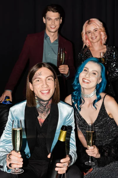 Tätowierter Mann mit Champagnerflasche blickt in die Kamera, während er Weihnachten mit nichtbinären Freunden auf schwarzem Hintergrund feiert — Stockfoto