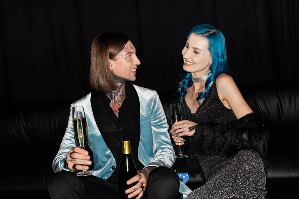 Stylish nonbinary friends sitting on couch with champagne and smiling at each other  on black background — Stock Photo